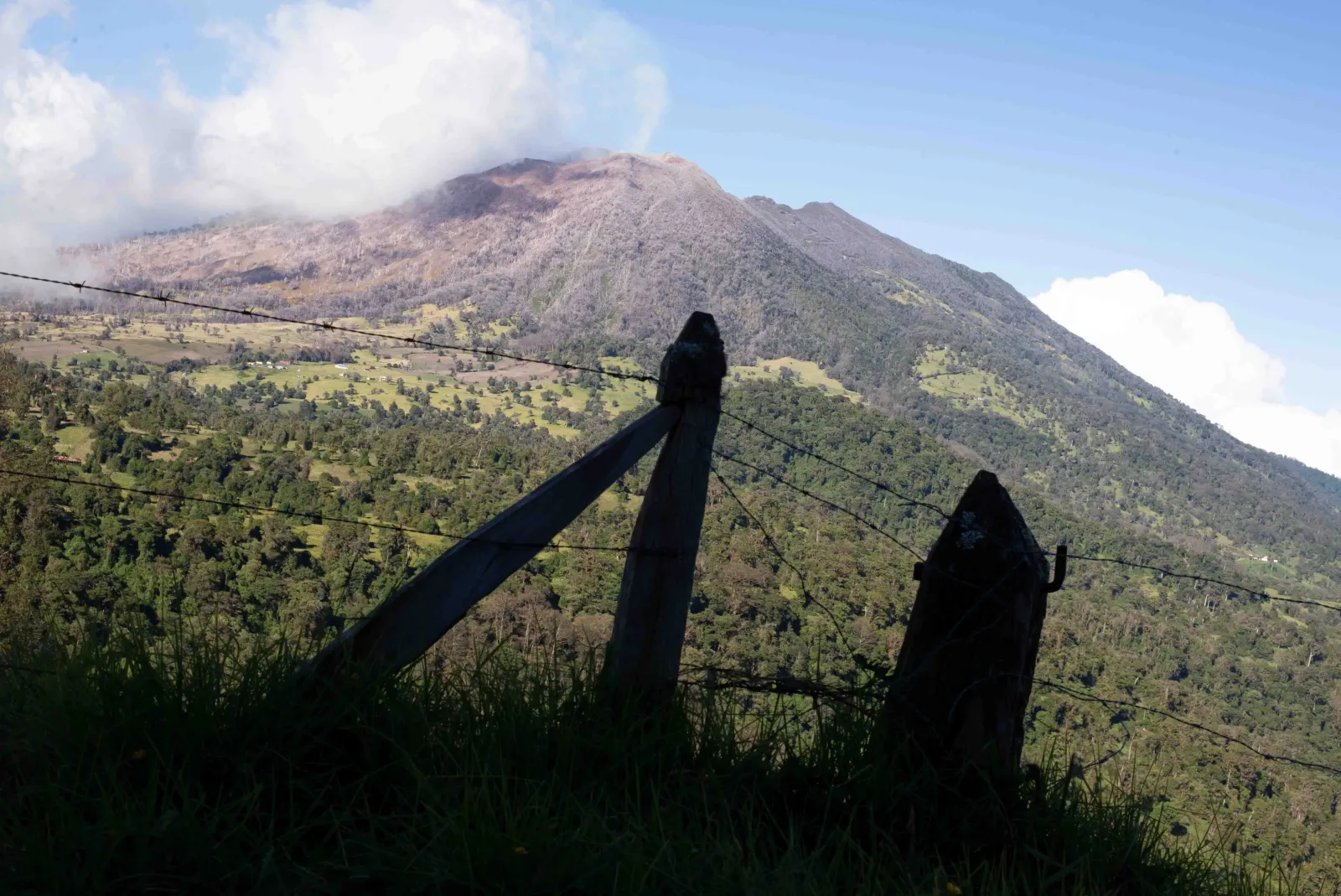 The Arenal volcano under cloud, seen across green pasture and a fence line